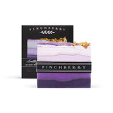 Square soap bar with varying shades of purple horizontal lines, going from dark on the bottom to lighter shades towards the top. The top of the bar has gold flecks sprinkled along it. In the background, the black packaging box is partially covered by the soap bar. Both the bar and the box have the Finchberry logo in white letters over a black background.