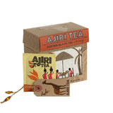 Brown Cardboard box with orange label and Kenyan artwork on the front against a white background. Yellow tea bag with similar artwork and tag is leaning against the box.