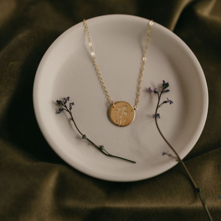 Gold circle floral stamped necklace with gold chain lays in a round white dish next to some lavender stems