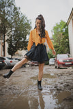 Woman wearing a yellow top, black skirt, and black roma boots splashes her feet in a muddy puddle
