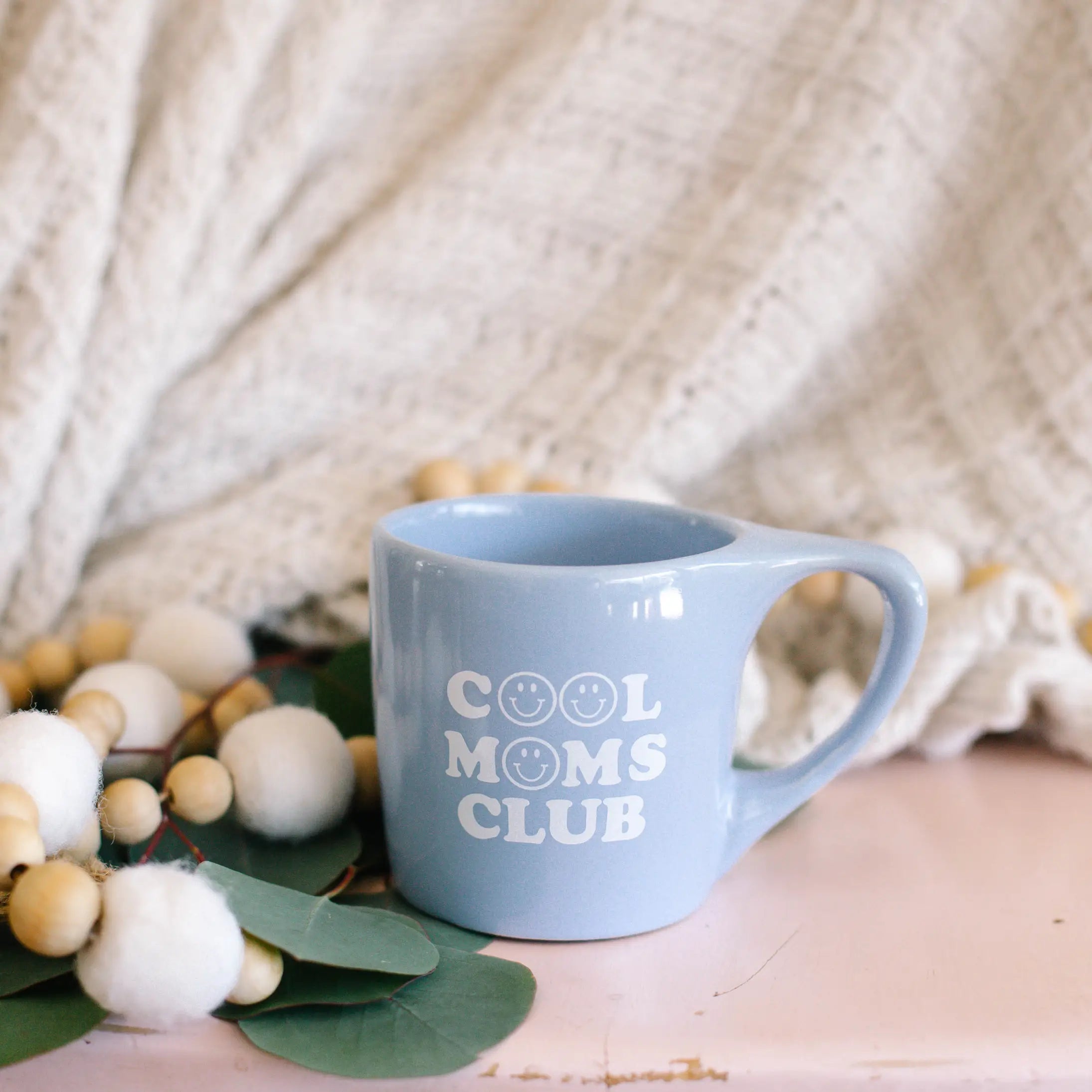 Cool Moms Club Ceramic Mug in Pastel Blue with Smiley Face from Rachel Allene with eucalyptus background