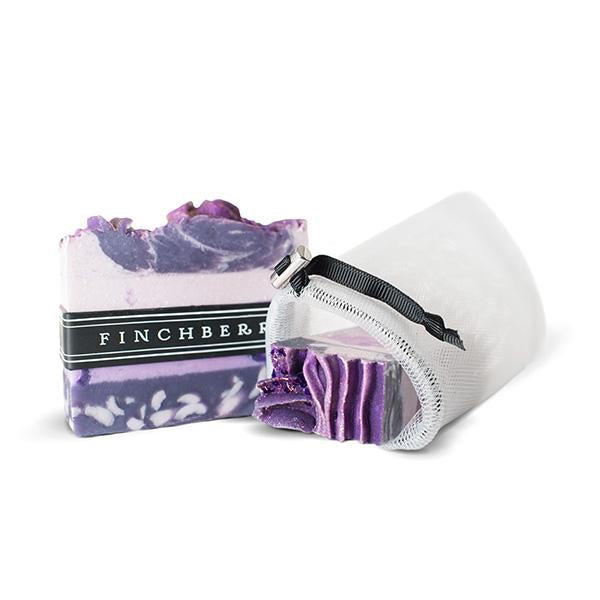 White mesh loofah pouch with black draw string laying on its side with a purple finchberry soap bar sitting next to it and part of the soap bar in the pouch. Against solid white backdrop