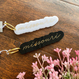 Two missouri acrylic keychains each with gold cursive font. One is white and one is black. There are pink florals over the wood floor surface