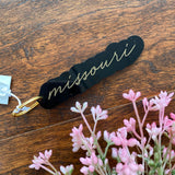 Black acrylic keychain with missouri written in gold cursive font. Sitting on wood surface with pink florals