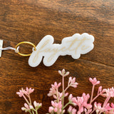 White acrylic keychain with fayette written in gold cursive font. Sitting on wood surface with pink greenery