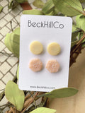 White beckhillco card sits over greenery while holding two pairs of stud earrings. One pair is yellow circles, another is blush hexagons. Both pairs are embossed with textured design