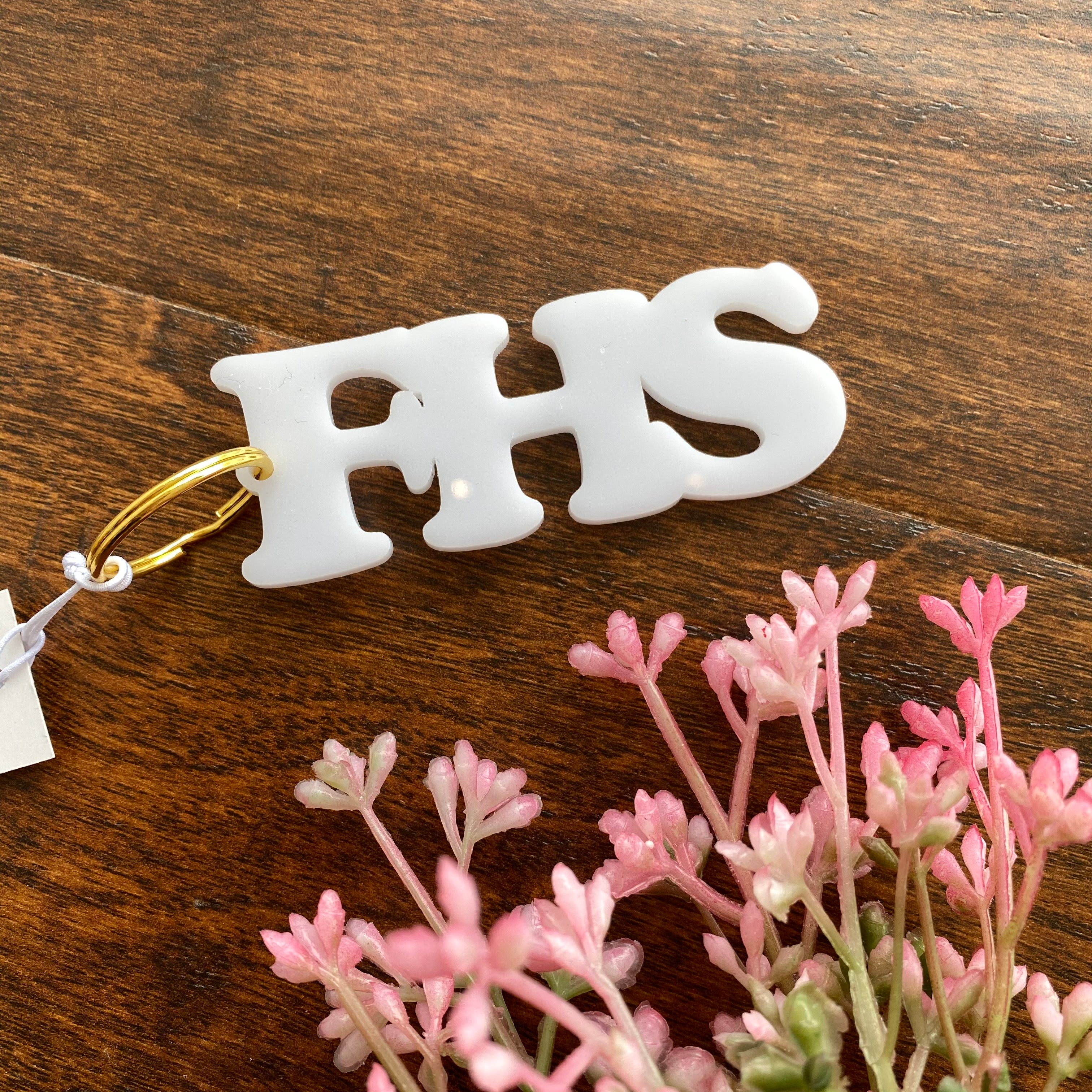 FHS initials make up white acrylic keychain in bold font. Wood surface with pink florals in the corner