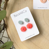 White beckhillco card sits on a wood stump while holding two pairs of earring studs. One is a vibrant coral colored hexagon shape, the other is a dusty blue sage oval shape. Both pairs are embossed with a textured detail