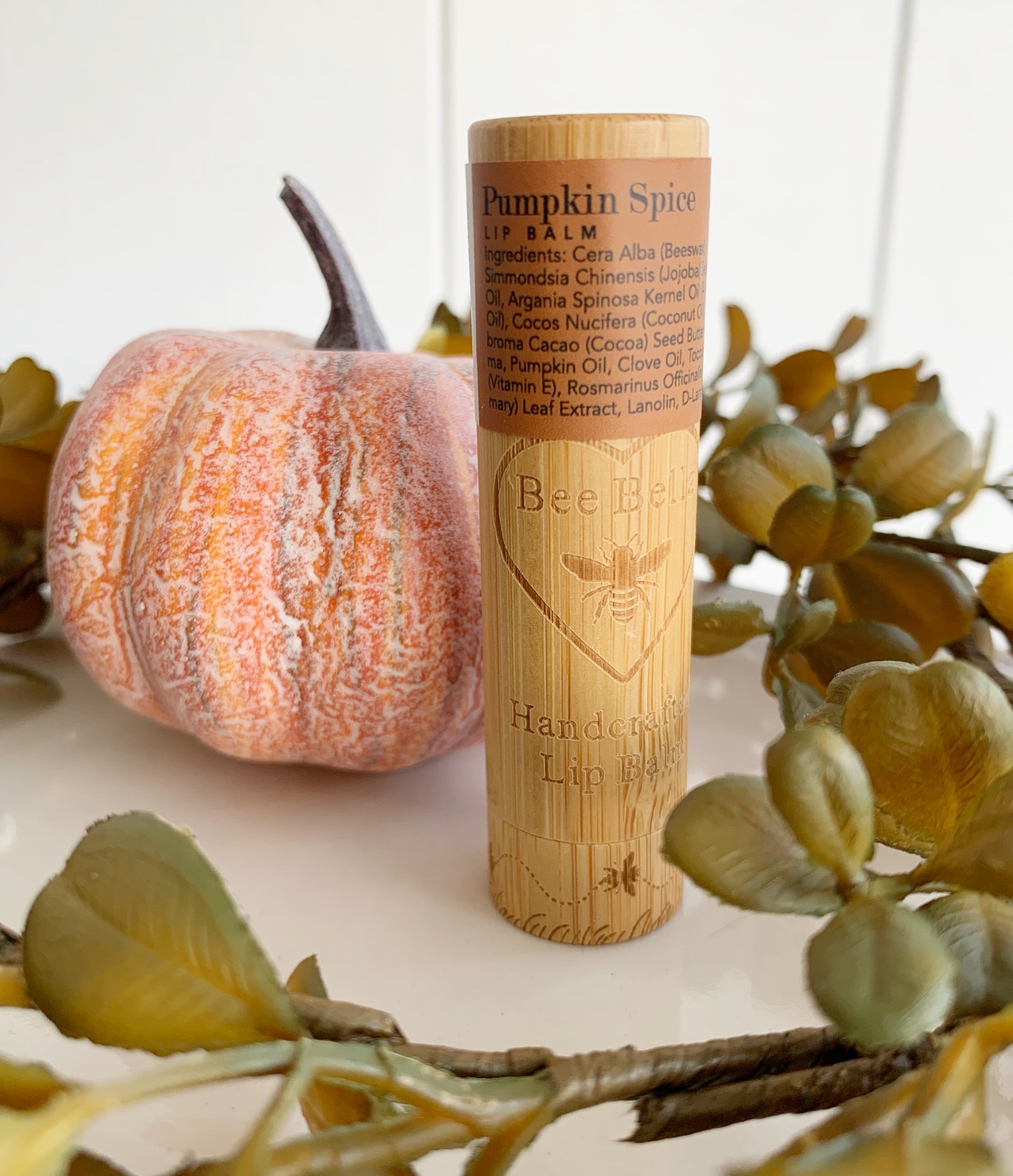 Wooden pumpkin spice lip balm tube with bee bella logo embossed and a brown flavor label at the top. Sitting upright surrounded by greenery and a pumpkin to the left.