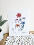 Art print of outline of a woman's face and arm as well as a floral arrangement with watercolor accents. Print sits on wooden tray filled with white pebbles against white wall