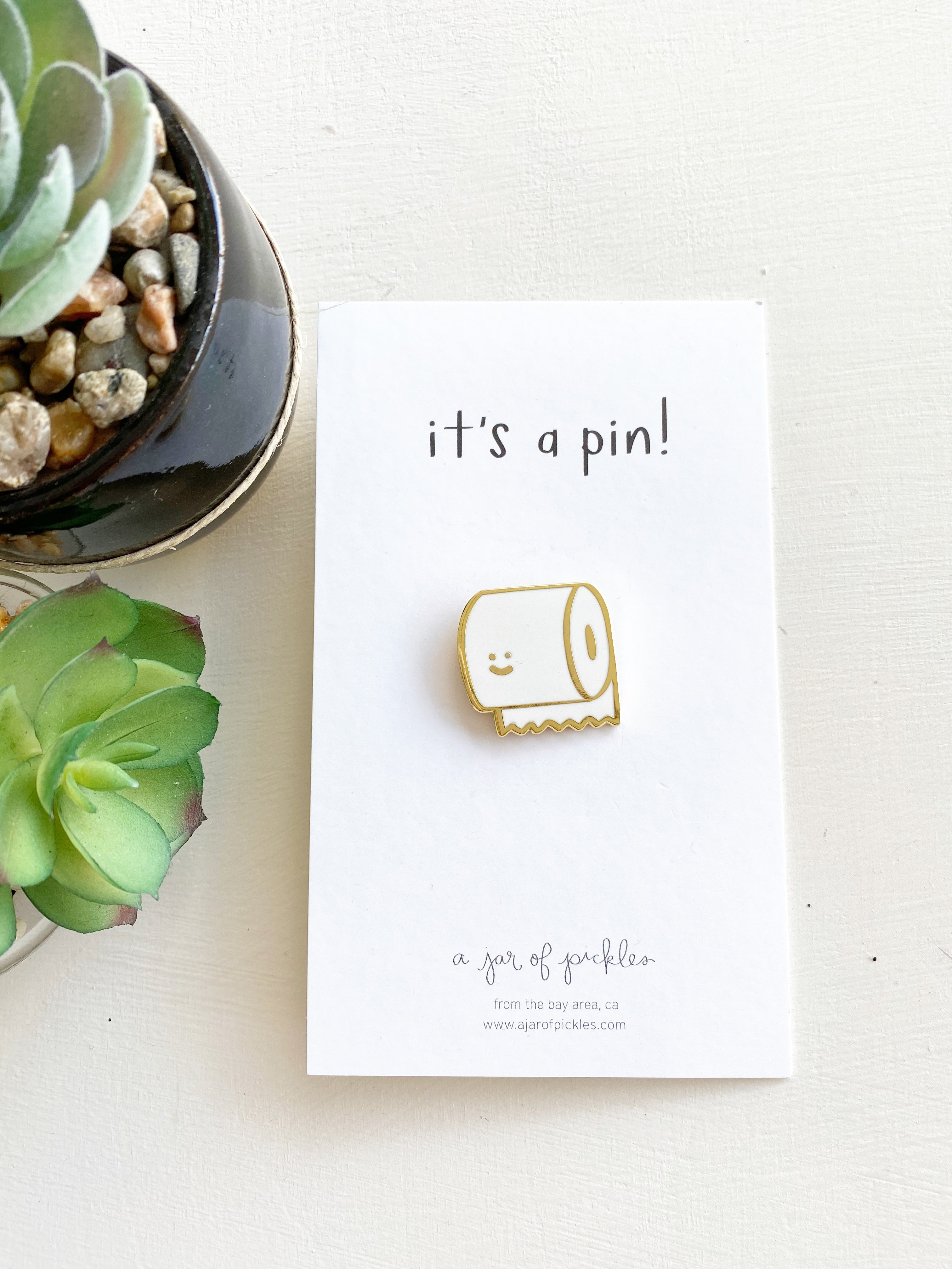 Gold and white toilet paper pin with a smiley face sits on a white card that reads ITS A PIN! over white surface next to succulents