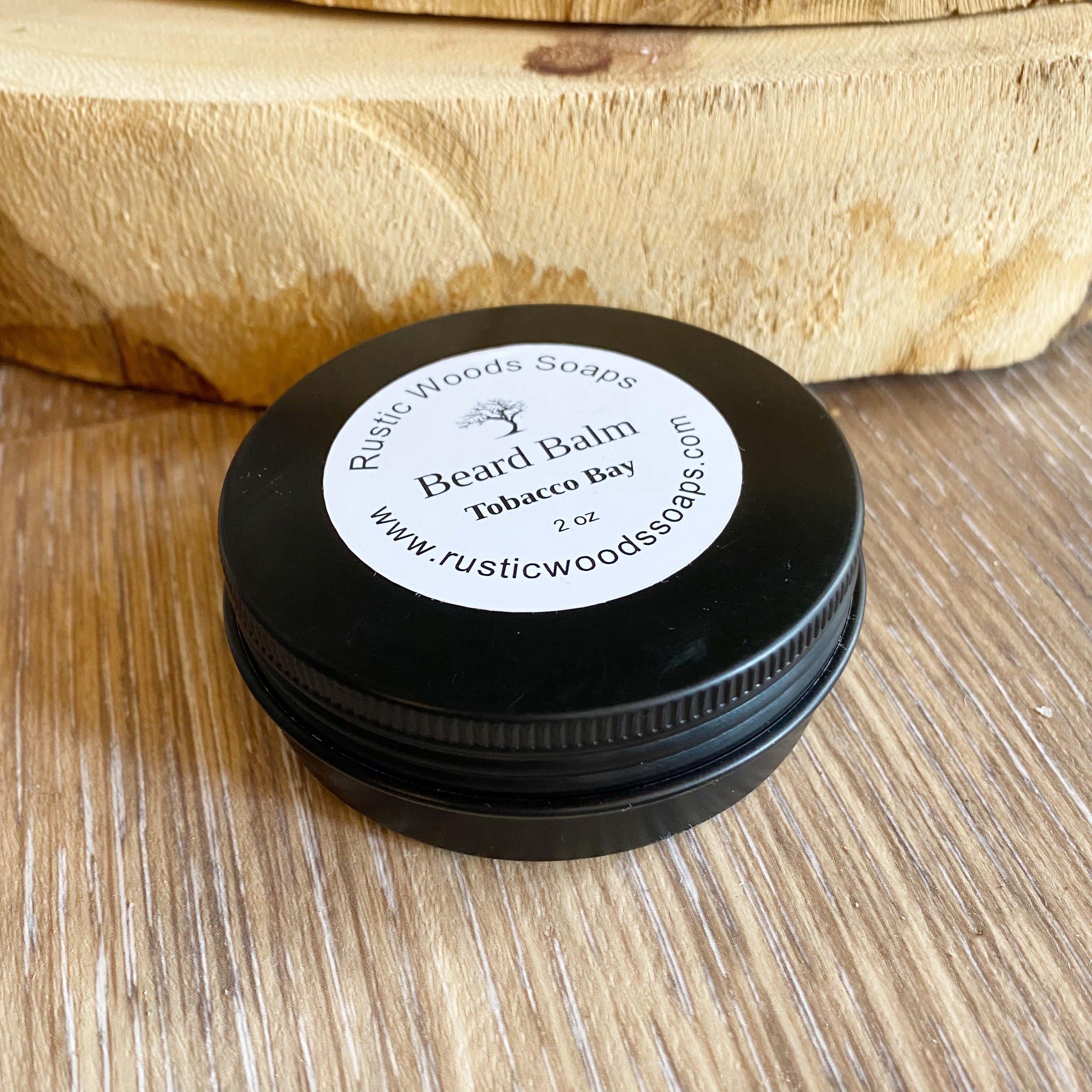 Black canister jar with white rustic woods soaps tobacco bay beard balm label lays on wood surface