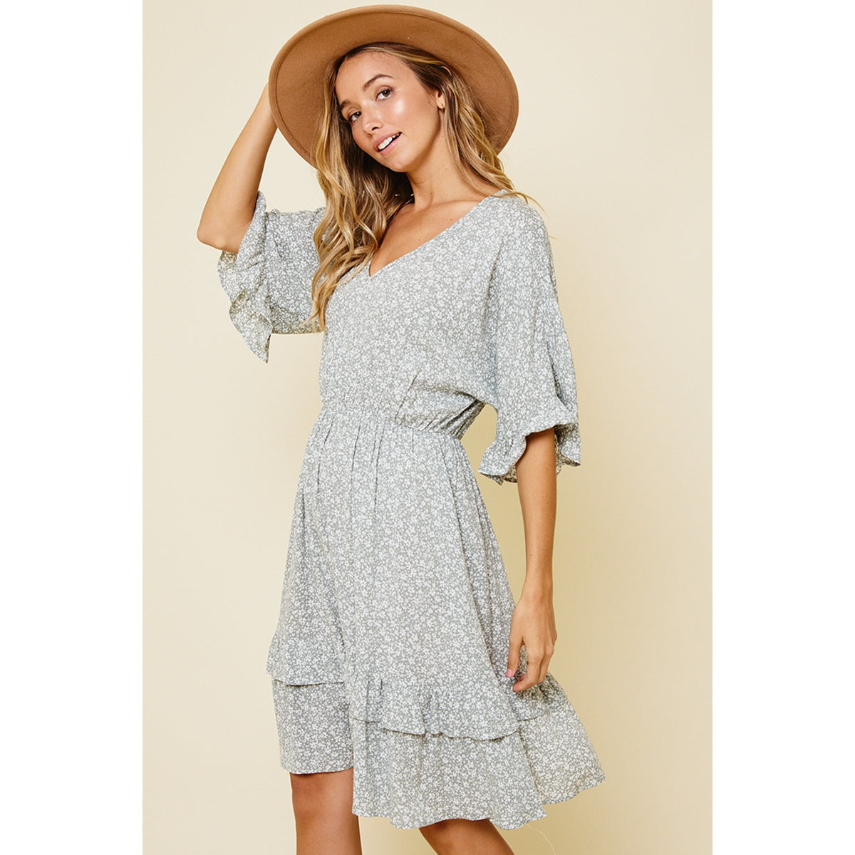 Side view of blonde model posing in a ruffle v-neck dress with a sage and white daisy floral print paired with a tan suede hat