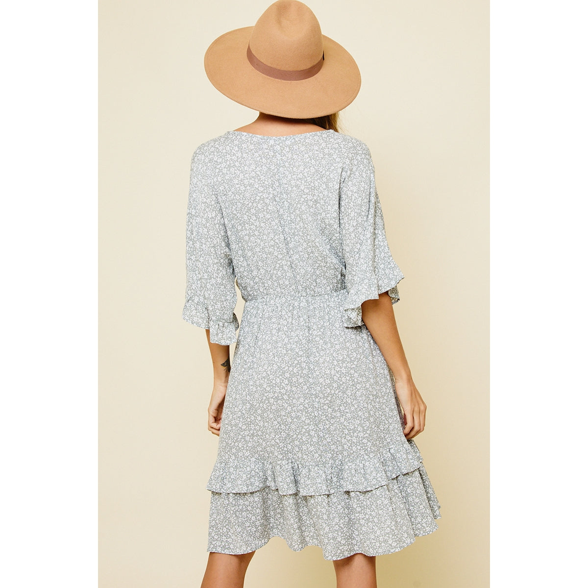 Back view of blonde model wearing a ruffle dress with a sage and white daisy floral print paired with a tan suede hat