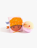 Cube box depicting maya angelou with birds sits on solid white backdrop next to pink and yellow bath bomb